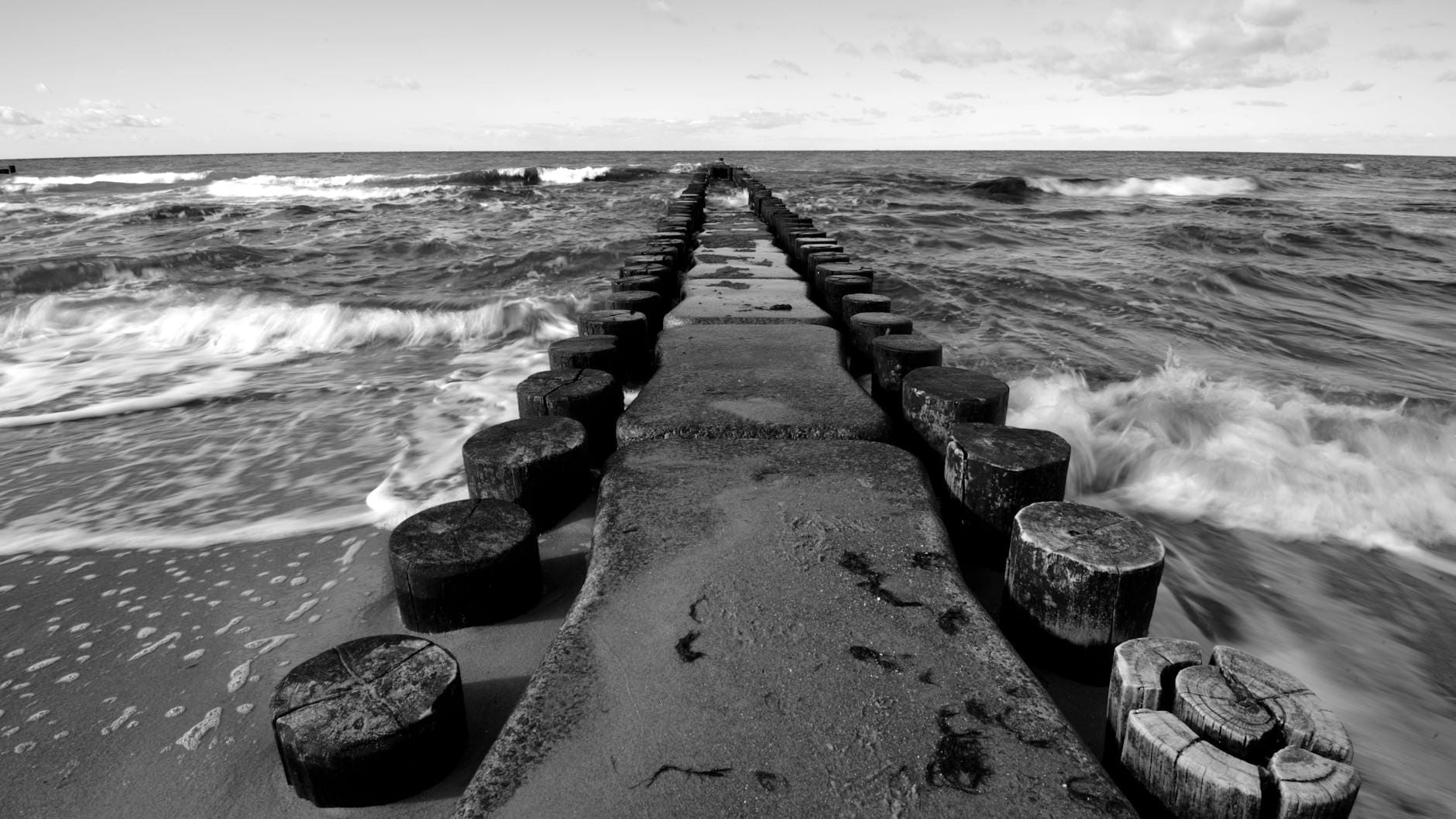 Czarno-białe fale Bałtyku rozbijające się o drewniany falochron
