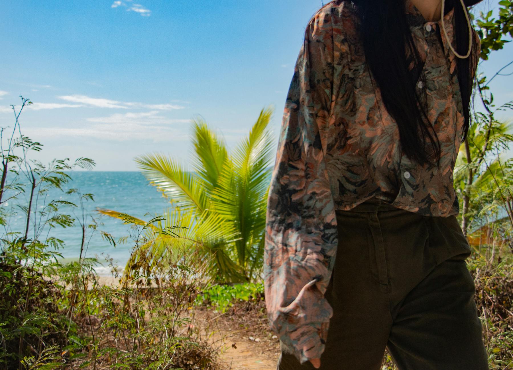 Kobieta w kwiecistej koszuli idzie brzegiem morza przy palmach